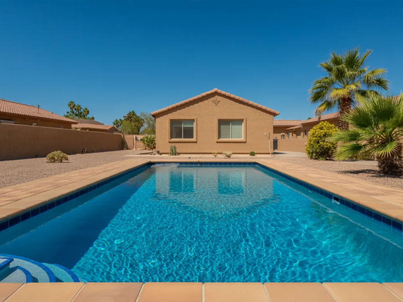 A rectangular in-ground swimming pool in a desert-style backyard with a single-story house, palm trees, and clear blue sky.